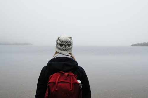 Woman wearing beanie, jacket and backpack looking out over a lake
