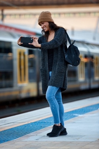 Woman wearing beanie taking photo with phone at train station - Australian Stock Image