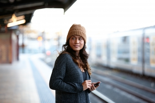 Woman wearing beanie waiting at train station with mobile phone - Australian Stock Image