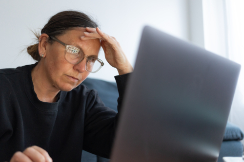 Woman wearing black top and eyeglasses looking serious while working on laptop at home - Australian Stock Image