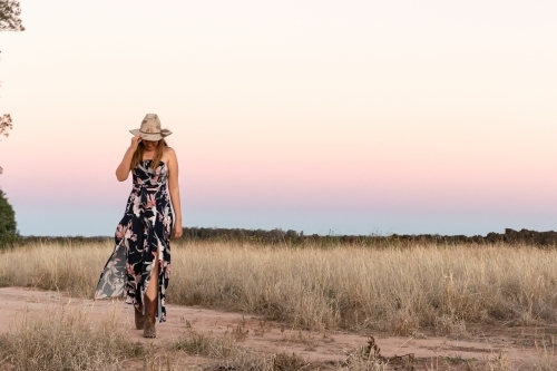 Woman wearing floral dress cowboy boots and hat while walking on a dirt road in rural area - Australian Stock Image