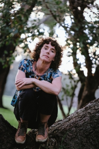 woman with curly hair and casually dressed sitting on tree trunk - Australian Stock Image