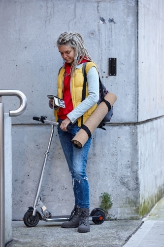 Woman with dreadlocks on electric scooter going to yoga class with phone - Australian Stock Image