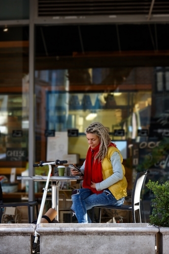 Woman with dreadlocks texting at cafe with electric scooter - Australian Stock Image