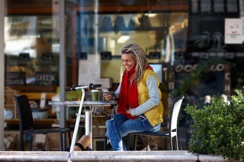 Woman with dreadlocks using her mobile phone at cafe with electric scooter - Australian Stock Image