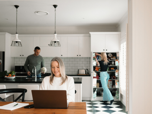 Woman working from home on her laptop at wooden table while husband works in the kitchen - Australian Stock Image