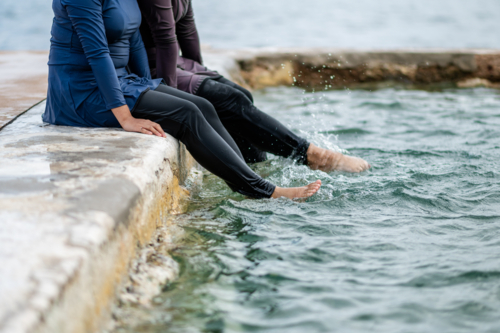Women in modest swimwear sitting on stone platform kicking their legs in the tidal pool - Australian Stock Image