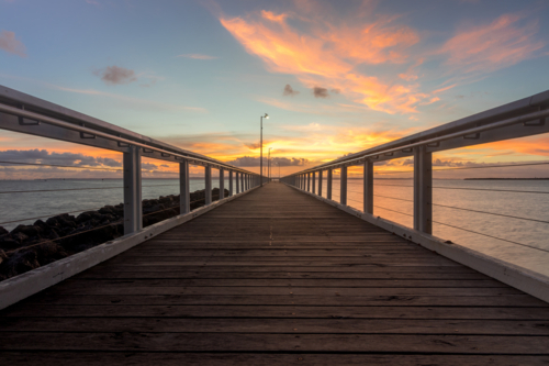 Wooden jetty stretching over water with cloudy sunrise sky - Australian Stock Image