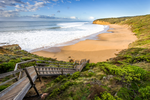 Wooden staircase leading down to a sandy beach with rolling waves and rugged cliffs - Australian Stock Image