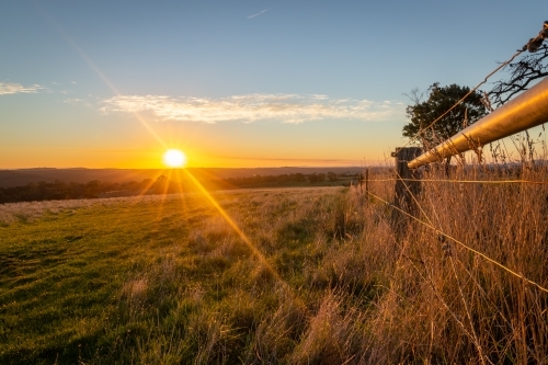 Yellow Sun setting in the distance over hills and paddocks with a farm fence framing the image - Australian Stock Image