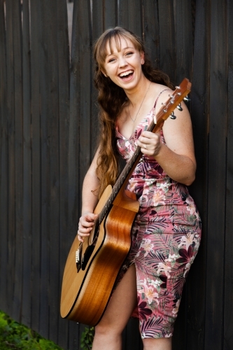 Young adult guitar player playing instrument outside leaning on black wooden fence - Australian Stock Image