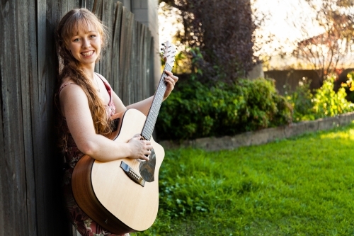 Young adult guitar player playing instrument outside leaning on black wooden fence - Australian Stock Image