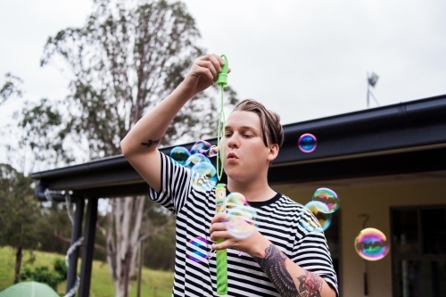 Young adult male with tattoos blowing multiple bubbles outside.