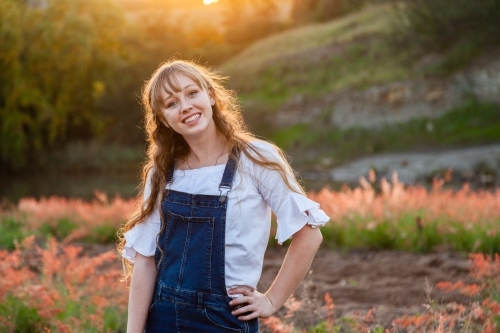 Young adult standing outside by river at sunset in overalls - Australian Stock Image