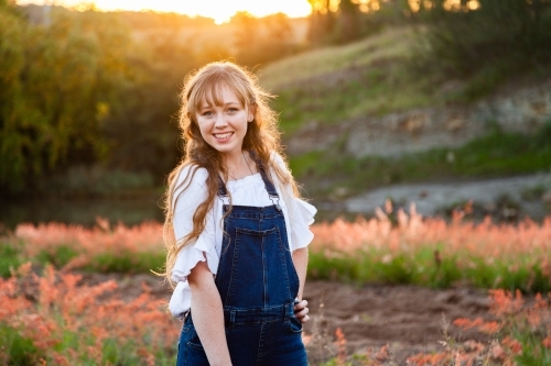 Young adult standing outside by river at sunset in overalls - Australian Stock Image