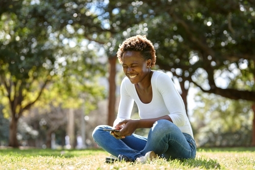 Young African woman laughing in sunshine at park with mobile phone - Australian Stock Image