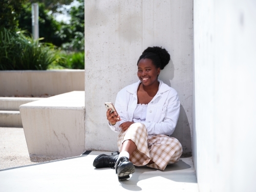 Young African woman using a phone sitting against a concrete wall - Australian Stock Image