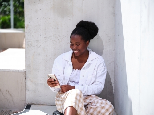 Young African woman using a phone sitting against a concrete wall - Australian Stock Image