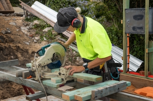 Young apprentice builder cutting length of timber with drop saw on residential construction site - Australian Stock Image