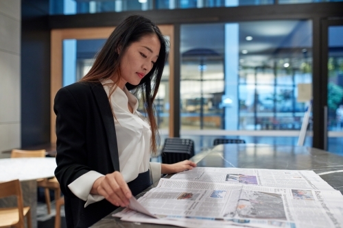 Young Asian business woman drinking coffee and reading newspaper - Australian Stock Image