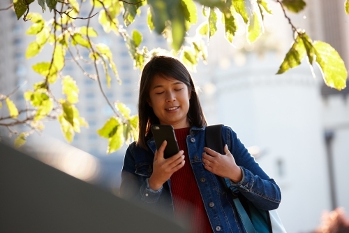 Young Asian female checking mobile phone under tree - Australian Stock Image
