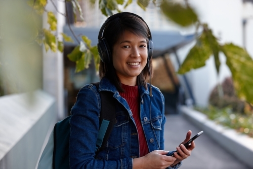 Young Asian female listening to music with headphones under tree - Australian Stock Image