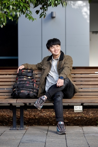 Young Asian man smiling sitting on park bench whilst listening to wireless headphones