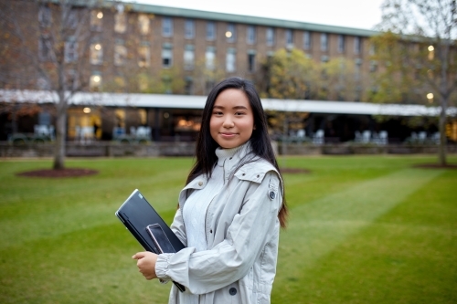 Young Asian student holding her laptop on lawn at university campus
