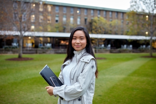 Young Asian student holding her laptop on lawn at university campus - Australian Stock Image