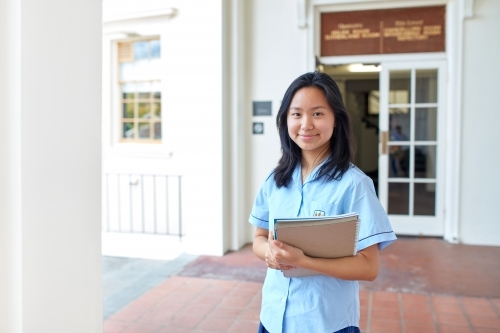 Young Asian student in front of school building holding books - Australian Stock Image