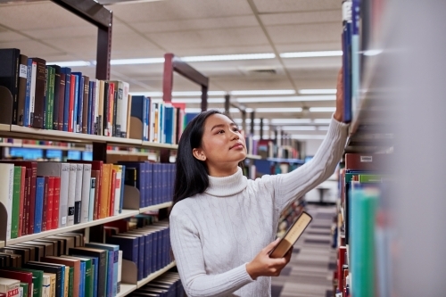 Young Asian student standing in aisle at university library - Australian Stock Image