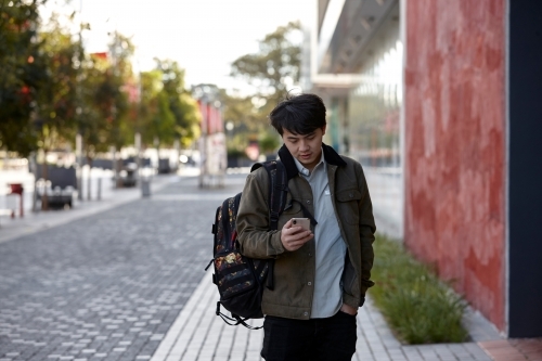 Young Asian university student walking and checking phone messages on-campus - Australian Stock Image