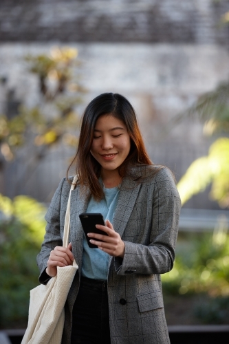 Young Asian woman with shopping bag and phone outdoors at enclave - Australian Stock Image