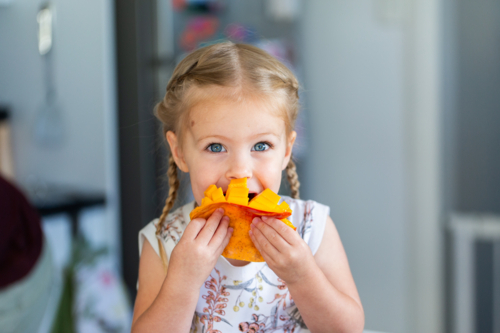 Young Australian toddler kid eating fresh mango fruit in kitchen at home during summer - Australian Stock Image