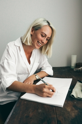 Young blonde woman laughing as she sits at a desk drawing in a sketch book - Australian Stock Image