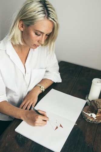 Young blonde woman sitting at desk drawing in a sketch book - Australian Stock Image