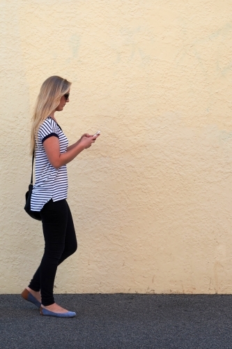 Young blonde woman walking and texting on mobile phone - Australian Stock Image