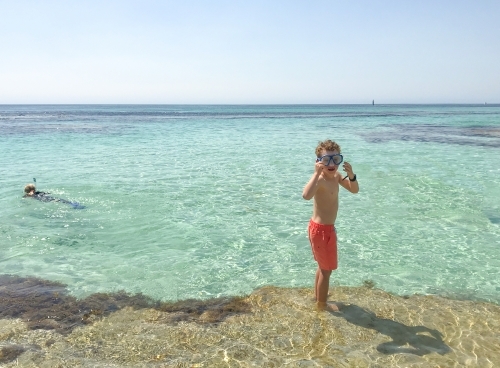 young boy adjusting mask on edge of clear lagoon standing on reef in orange board shorts