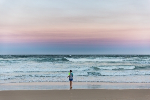 Young boy alone on beach at sunset looking at waves - Australian Stock Image