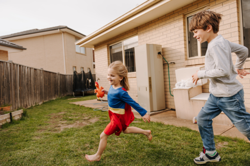 Young boy chasing after the little girl in superhero costume in the yard. - Australian Stock Image