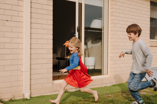 Young boy chasing after the little girl in superhero costume in the yard. - Australian Stock Image