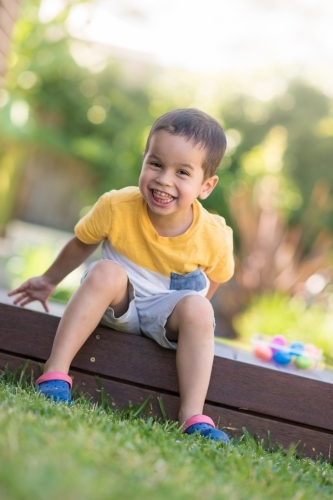 Young boy happily sitting on deck next to his eggs after easter egg hunt. - Australian Stock Image