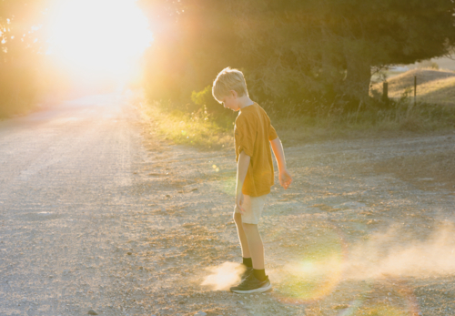 young boy kicking dirt on side of country road at sunset - Australian Stock Image