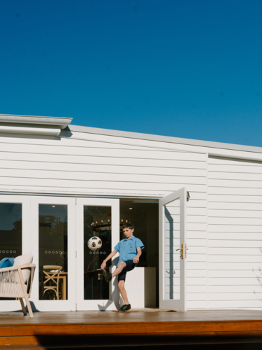 Young boy kicking soccer ball on garden deck of white weatherboard home - Australian Stock Image