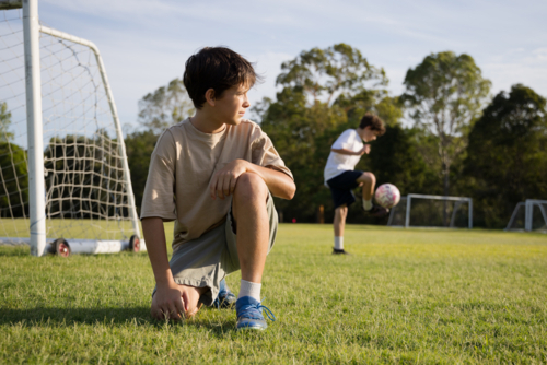 Young boy kneeling with one knee on grassy soccer field looking away - Australian Stock Image