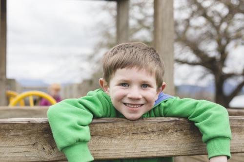 Young boy leaning on wooden railing on playground smiling - Australian Stock Image