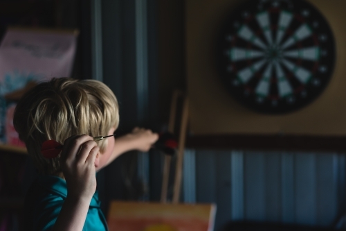 Young boy learning to play darts in shed - Australian Stock Image