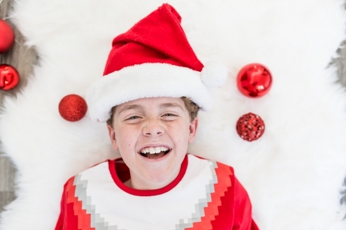 Young boy lying on rug with baubles wearing santa hat laughing at Christmas - Australian Stock Image
