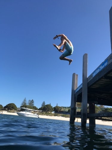 Young boy mid-air jumping of jetty With boat and beach in background - Australian Stock Image