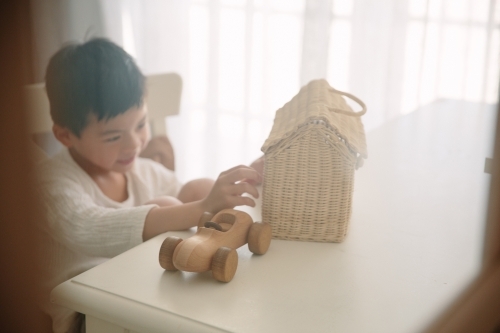 Young boy playing with a wooden toy car and rattan house bag while sitting on a table